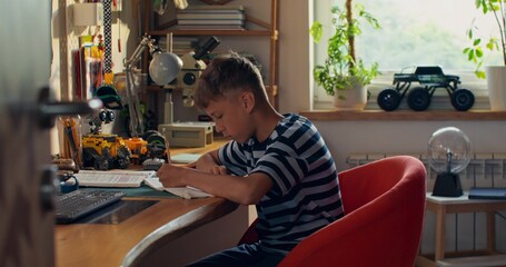 The boy is concentrating on doing his homework while sitting at the table at home