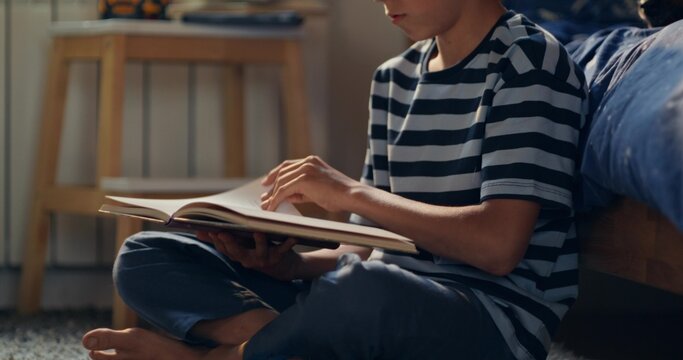 A boy is enthusiastically reading a book sitting on the floor in his room. His dog is on the bed next to him