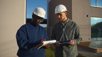Two men in construction helmets sign a contract standing outside the house