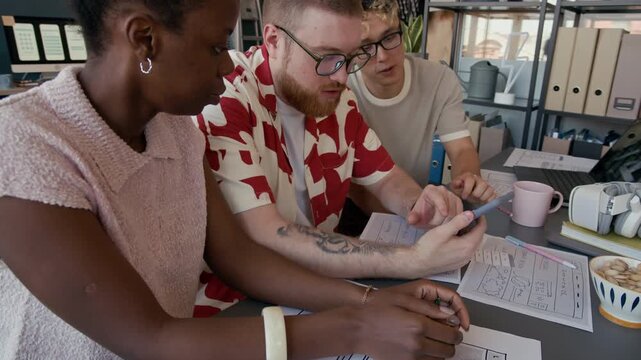 Diverse team of UI designers testing mobile app on smartphone developing user interface collaboratively while sitting at cluttered with papers desk together in modern office space
