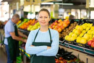 Positive middle-aged saleswoman standing in front of food stall in grocery store with large assortment