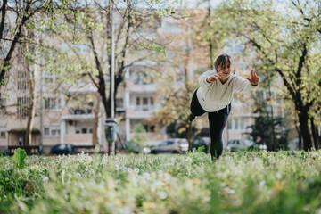 A woman in a white hoodie performs a balancing exercise on a grassy field in an urban park,...