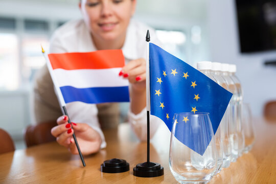 Young woman in business clothes puts flags of Netherlands and European Union on negotiating table in office