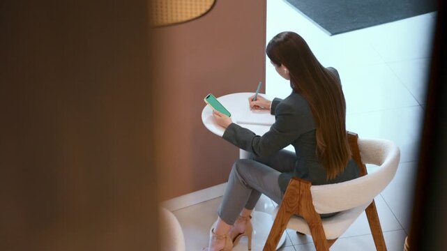 Businesswoman writing in notebook while holding phone in bright office lounge. Concept of business education, remote work, productivity, digital planning, women entrepreneurship.