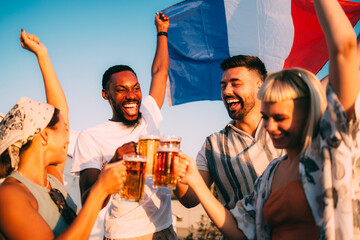 Happy multi-ethnic friends cheering with beer glasses and waving french flag at sunset
