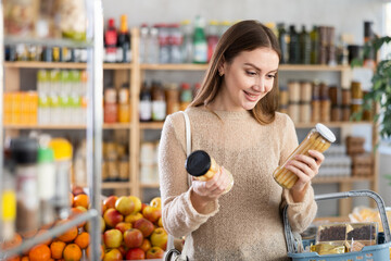 Casual young woman in cozy knitted sweater examining jars of marinated white asparagus in brine...