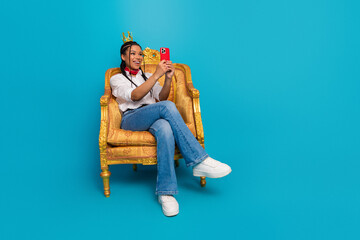 Young stylish woman sitting on ornate golden chair taking a selfie with a smartphone against a blue...