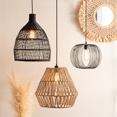 Boho chic inspired scene with black wicker dome pendant, black wire oval lamp and hexagonal rattan light, warm bulbs illuminating beige wall with woven round mirror and soft pampas grass accent.
