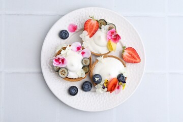Delicious dessert. Tasty tartlets with cream, berries and flowers on white tiled table, top view