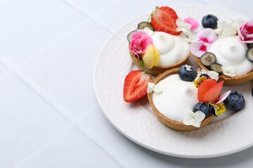 Delicious dessert. Tartlets with cream, berries and flowers on white tiled table, closeup. Space for text
