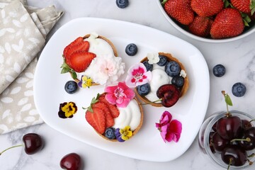 Sweet tartlets with berries and flowers on light marble table, flat lay. Delicious dessert