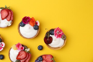 Sweet tartlets with berries and flowers on yellow background, flat lay with space for text. Delicious dessert