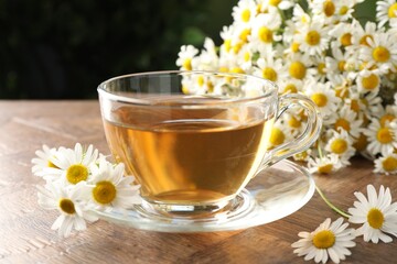 Aromatic tea in glass cup and chamomile flowers on wooden table, closeup