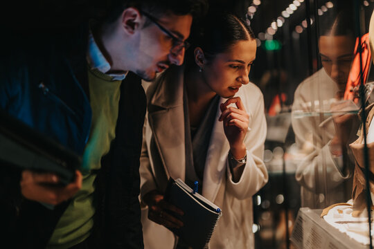 People closely examine artifacts displayed inside a museum showcase, displaying curiosity and intellectual engagement in the illuminated area, reflecting a theme of cultural exploration and education.