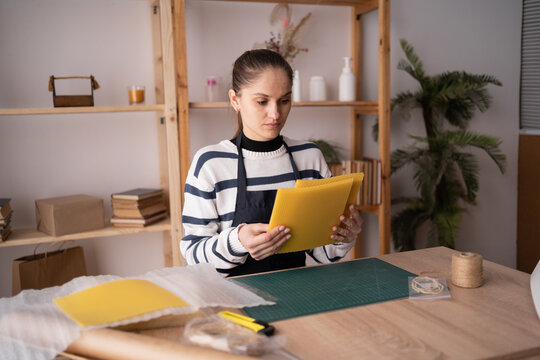 Hispanic brunette craftswoman in apron holding beeswax sheets making candles from natural wax sheet sitting at the table in workshop. - Powered by Adobe
