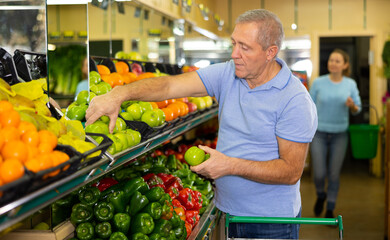 Attentive elderly man purchaser taking green apples from the counter in grocery store