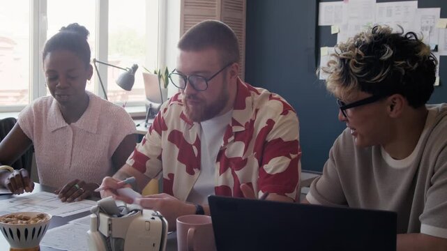 Cheerful Asian male UI designer collaborating with diverse creative team developing user interface of mobile app while meeting at desk with laptop and papers at modern office, slow motion