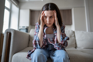 Frustrated depressed woman sitting on couch at home, hands pressed to temples, deep in anxious...