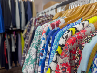 A colorful array of patterned clothing hangs on wooden hangers in a retail setting, with a blurred rack of more subdued garments in the background.