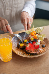 Close-up of a person cutting a slice of toast topped with fresh berries and cream using a fork and knife. Healthy breakfast or brunch scene, homemade food, natural lighting