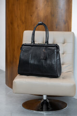 A vintage-style black leather duffel bag rests on a cream-colored tufted chair against a blurred wooden and grey background