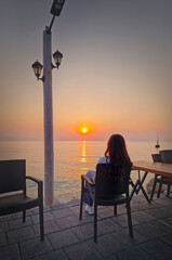 Rear view relaxed young woman sits on the waterfront watching the sunrise over the sea in Kemer, Turkey. Peaceful ambiance seaside with female tourist silhouetted against the vibrant dawn