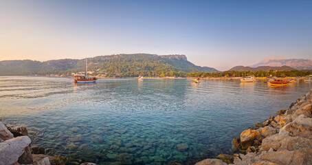 Morning light bathes the Moonlight beach in Kemer, Turkey. Boats rest in the calm bay with turquoise Mediterranean sea water. Lush green hills with palm trees seen at horizon