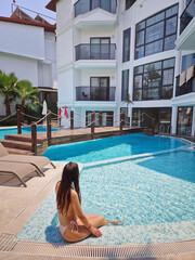 Beautiful woman in a pink bikini sits relaxed by a resort pool, gently splashing the water. The serene setting features a white hotel building, and yard with palm trees, and a wooden bridge
