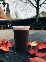 A cup of coffee sits on a table surrounded by fallen maple leaves in an autumn park