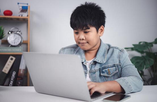 Young Asian boy focused and engaged with laptop at desk, ideal for representing online education, digital literacy, and children's interaction with technology in a home learning environment.