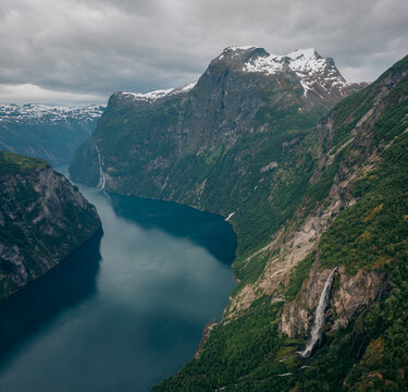 Aerial view of Geirangerfjord's dark waters winding between towering green cliffs and cascading waterfalls under a brooding sky, Stranda Municipality, M&Atilde;&cedil;re og Romsdal, Norway.