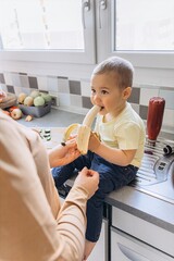 Toddler sits on a kitchen counter eating a banana, with a parent standing nearby. Bright morning...