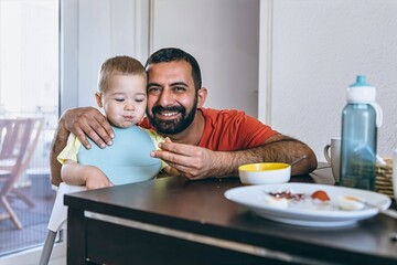 Father feeds his young son sitting in a high chair during breakfast. Morning family moment in the...