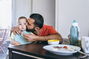 Father lovingly kisses his toddler on the cheek during breakfast time at home. A warm and emotional...