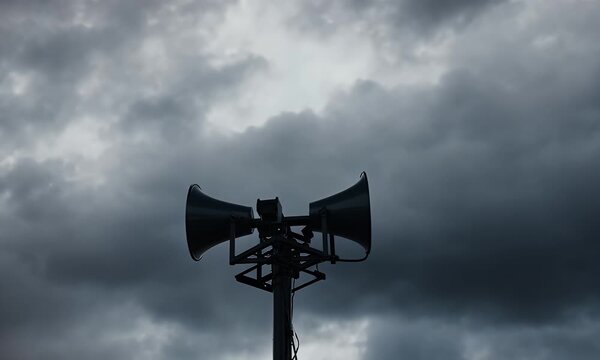 Silhouetted loudspeakers atop a tower against a moody, overcast sky, hinting at an impending storm