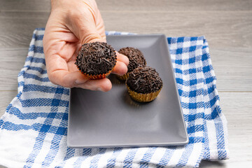 Hand holding a brigadeiro in a gold wrapper, with more chocolate truffles on a gray plate and blue...