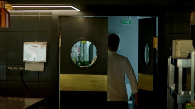 TRACKING Caucasian male chef walking out of commercial restaurant kitchen through the swinging doors. Shot with 2x anamorphic lens