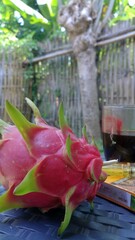 A whole dragon fruit with pink and green skin sits on an outdoor table, with a blurred garden background suggesting a relaxed tropical setting in Jember, East Java, Indonesia
