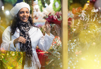 Hispanic woman standing near outdoor floral shop at christmas fair, touching leaves of bouquet and...