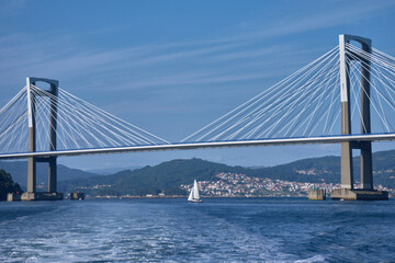Sea wake in the foreground as a boat moves away, leaving Rande Bridge behind in the Vigo Estuary on a clear day