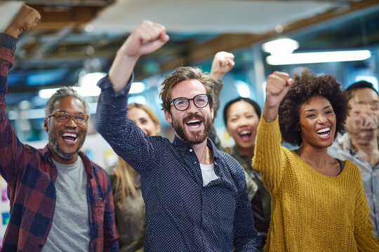 Diverse group celebrates success. Team members cheer with fists raised. Positive, energetic mood. Good for marketing, corporate, or editorial use.