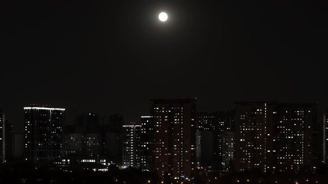 Time lapse. Rising moon over a big city at night. Hundreds of lights come on and go out in houses