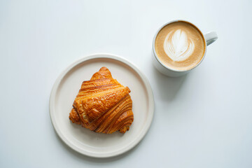 A golden brown croissant on a round plate next to a cup of coffee with latte art on a white background.
