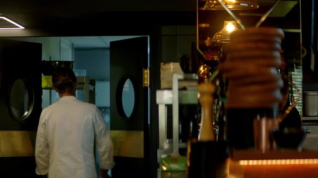 TRACKING Caucasian male chef walking out of commercial restaurant kitchen through the swinging doors. Shot with 2x anamorphic lens