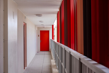 Couloir extérieur d’un bâtiment moderne avec, au fond, une porte rouge fermée et des barres en...