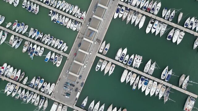 Aerial view of a marina filled with rows of white boats docked in calm teal waters, bisected by a pier with parked cars, Calaginesta, Spain.