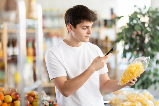 Before buying potato chips, young man scans barcode QR code on label of chips package, follows link and studies information for consumer in supermarket application - Powered by Adobe