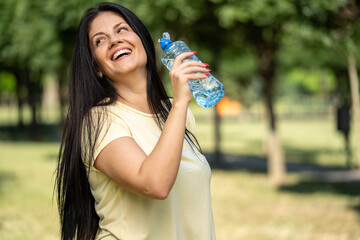Fit woman drinking water during morning workout in park, refreshing and hydrating body while...