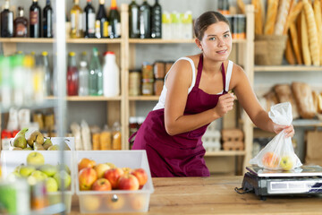 Young female seller weighing apples in plastic bag in grocery store