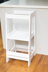 A sturdy white wooden step stool, designed for kitchen use, stands on a wooden floor against a...